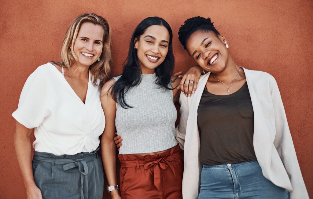 confident women smiling lingerie lifestyle milwaukee » milwaukeelingerie.com A group of three confident, smiling women standing close together against a rust-colored wall—used to reflect community, body positivity, and personal style in Milwaukee.